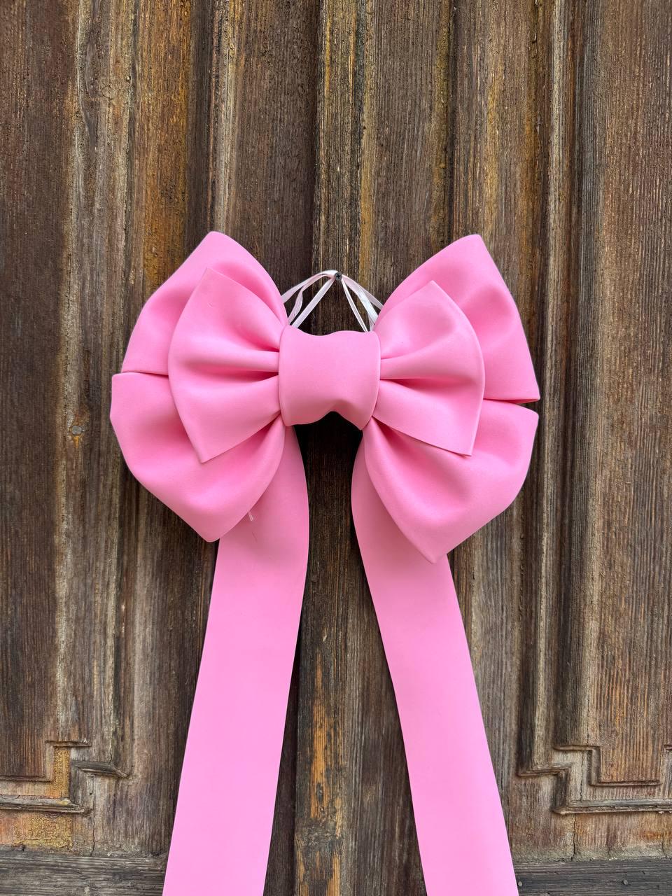 May include: A large, bright pink bow with long, flowing ribbons. The bow is made of a smooth, satin-like material and is suspended against a weathered, wooden background. The bow's design features a central knot and symmetrical loops.