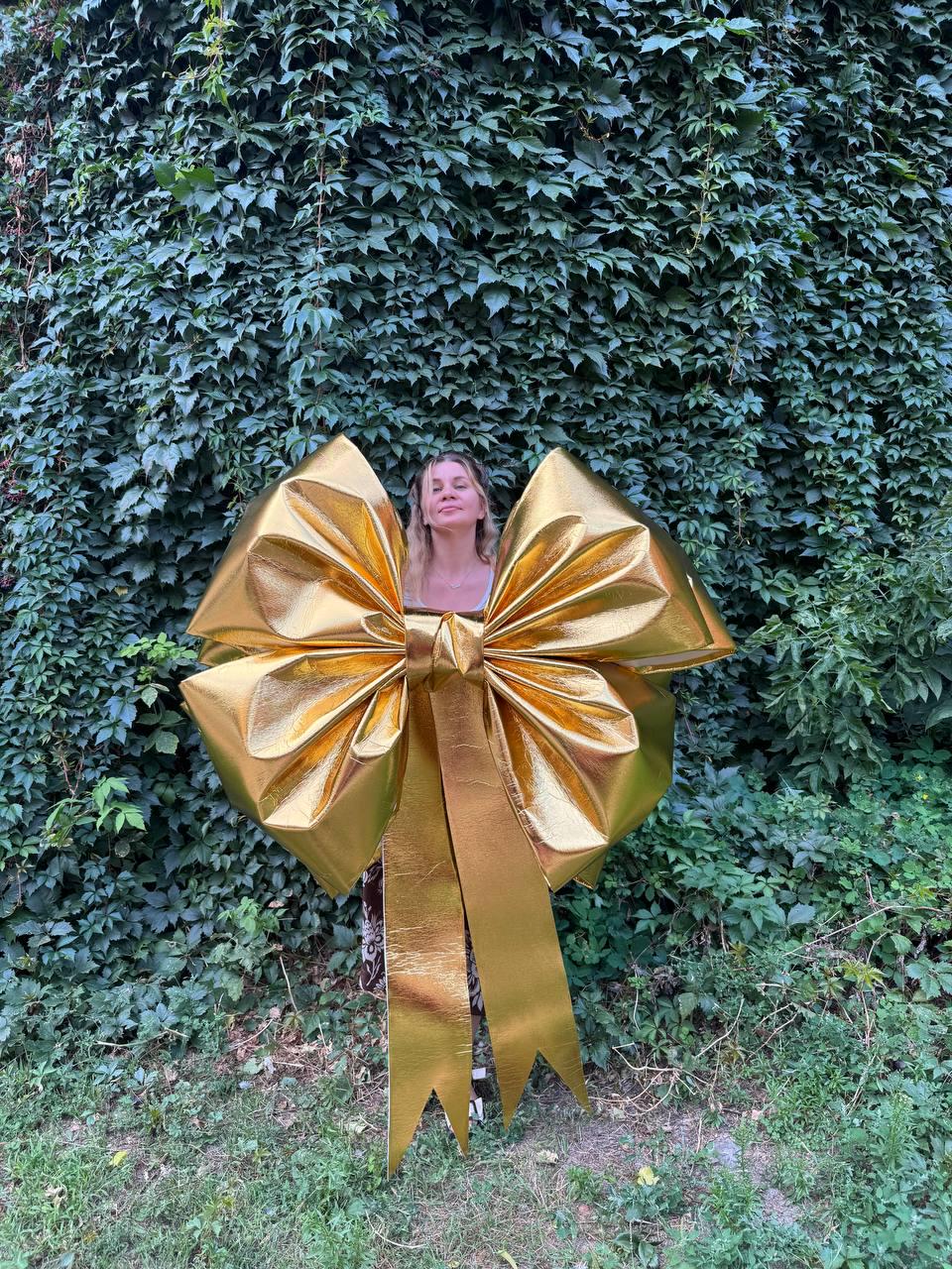 May include: A large, shiny gold bow with long, pointed streamers. The bow is the focal point, set against a backdrop of lush green foliage. The bow's metallic surface reflects light, creating a festive and eye-catching display.