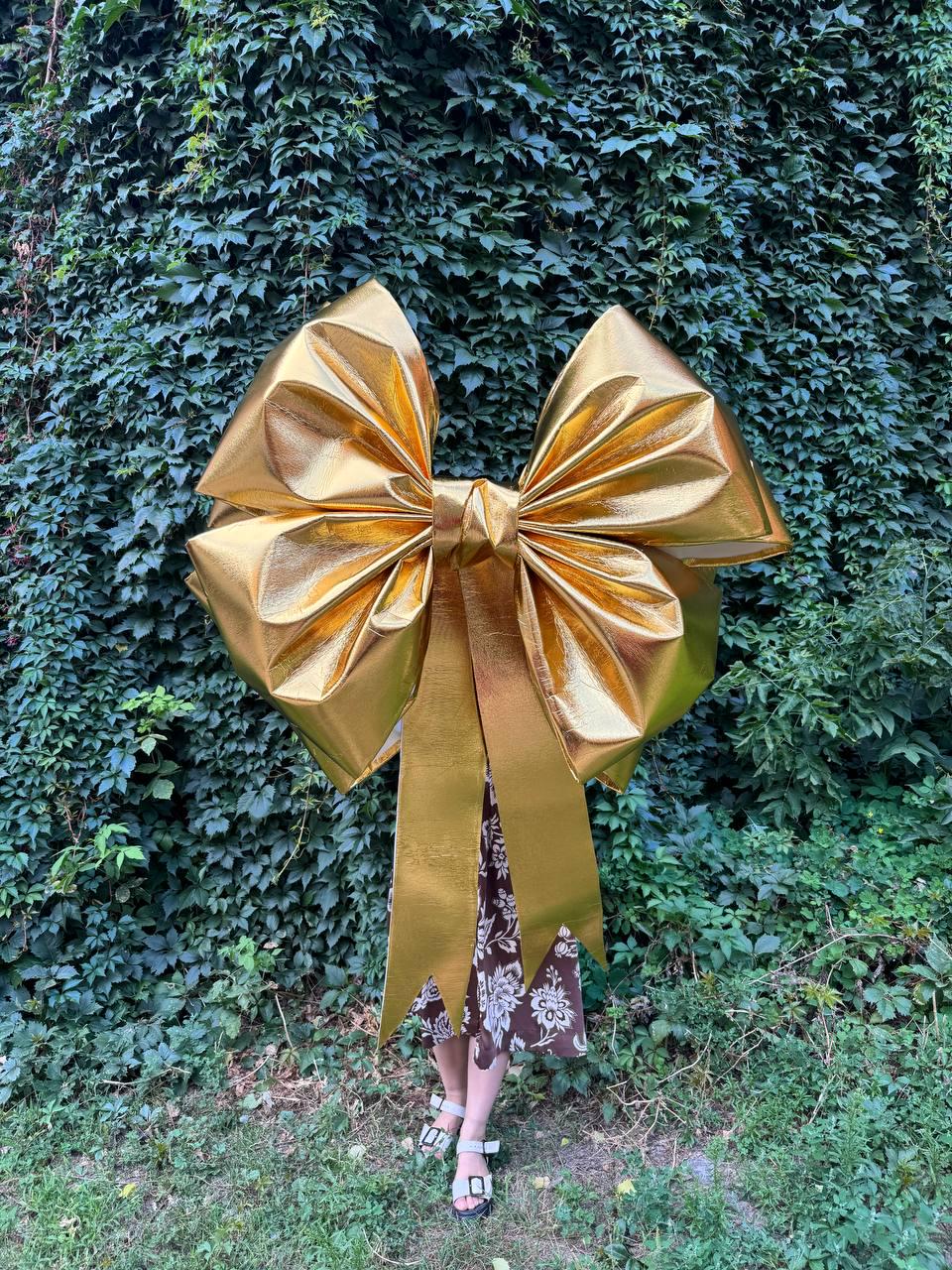 May include: A large, golden bow with long streamers. The bow is made of a shiny, metallic material and is positioned against a backdrop of green foliage. The bow's size suggests it is for decoration.