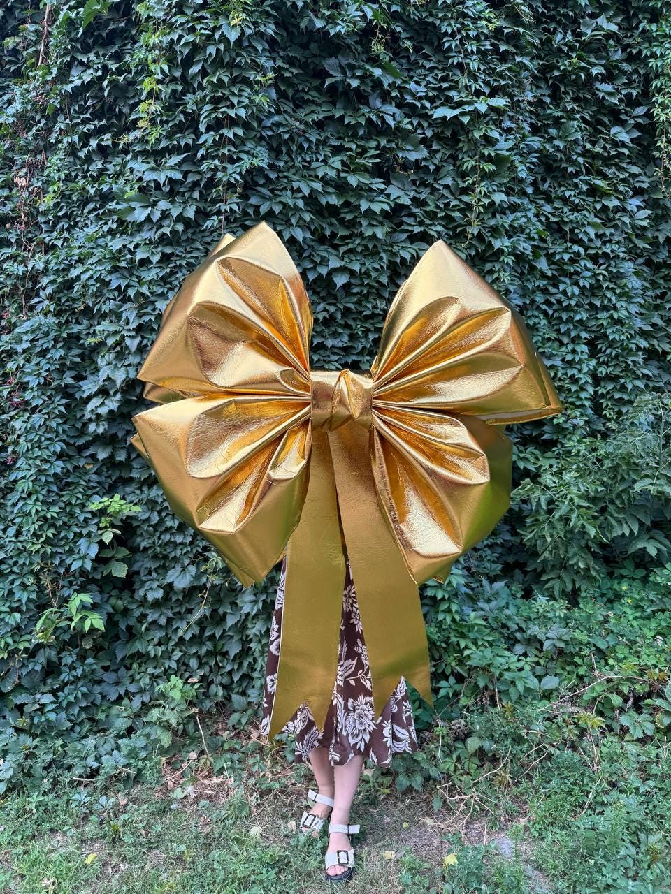 May include: A large, gold-colored bow with long, flowing ribbons. The bow is made of a shiny material and is positioned in front of a green, leafy background. The bow's size suggests it could be used for a large gift or decoration.