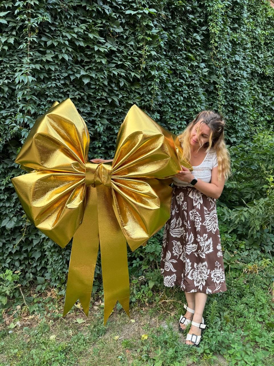May include: A large, golden bow with long, flowing ribbons. The bow is made of a shiny, metallic material and is positioned against a backdrop of green foliage. The bow is a decorative element, suitable for events or gift wrapping.