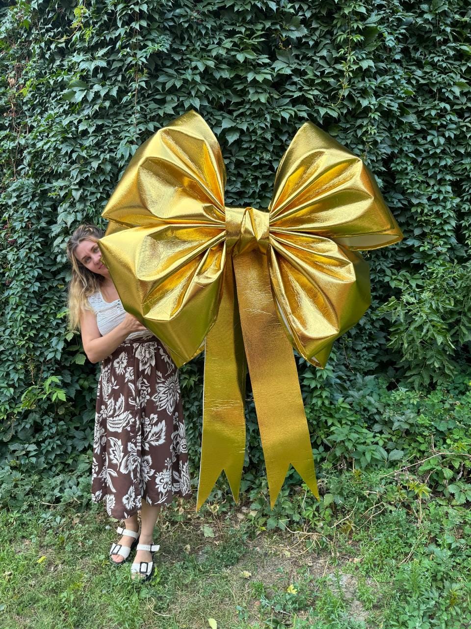 May include: A large, golden bow with long, pointed streamers. The bow is made of a shiny, metallic material and is positioned in front of a wall of green foliage. The bow is a decorative element, possibly for a gift or event.
