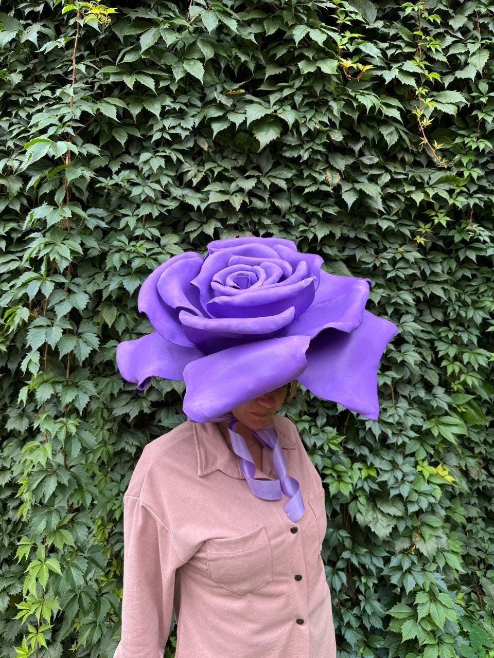 May include: A large, purple rose-shaped hat with a ribbon tie. The hat is worn by a person wearing a pink button-down shirt. The background is a wall of green leaves.