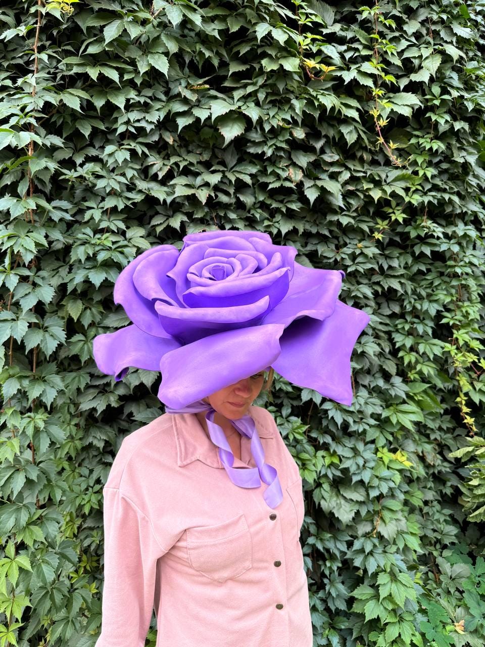 May include: A large, purple rose-shaped hat. The hat is worn over a pink button-down shirt. The background is a wall of green leaves. The hat's petals are detailed and the color is a vibrant purple.