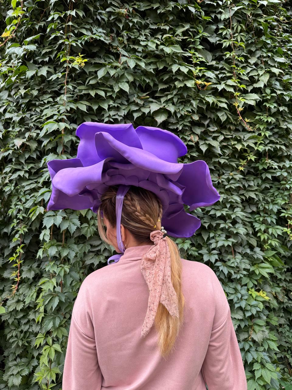 May include: A large, purple flower-shaped hat. The hat is worn by a person with blonde hair, a pink sweater, and a pink lace ribbon in their hair. The background is a wall of green leaves.