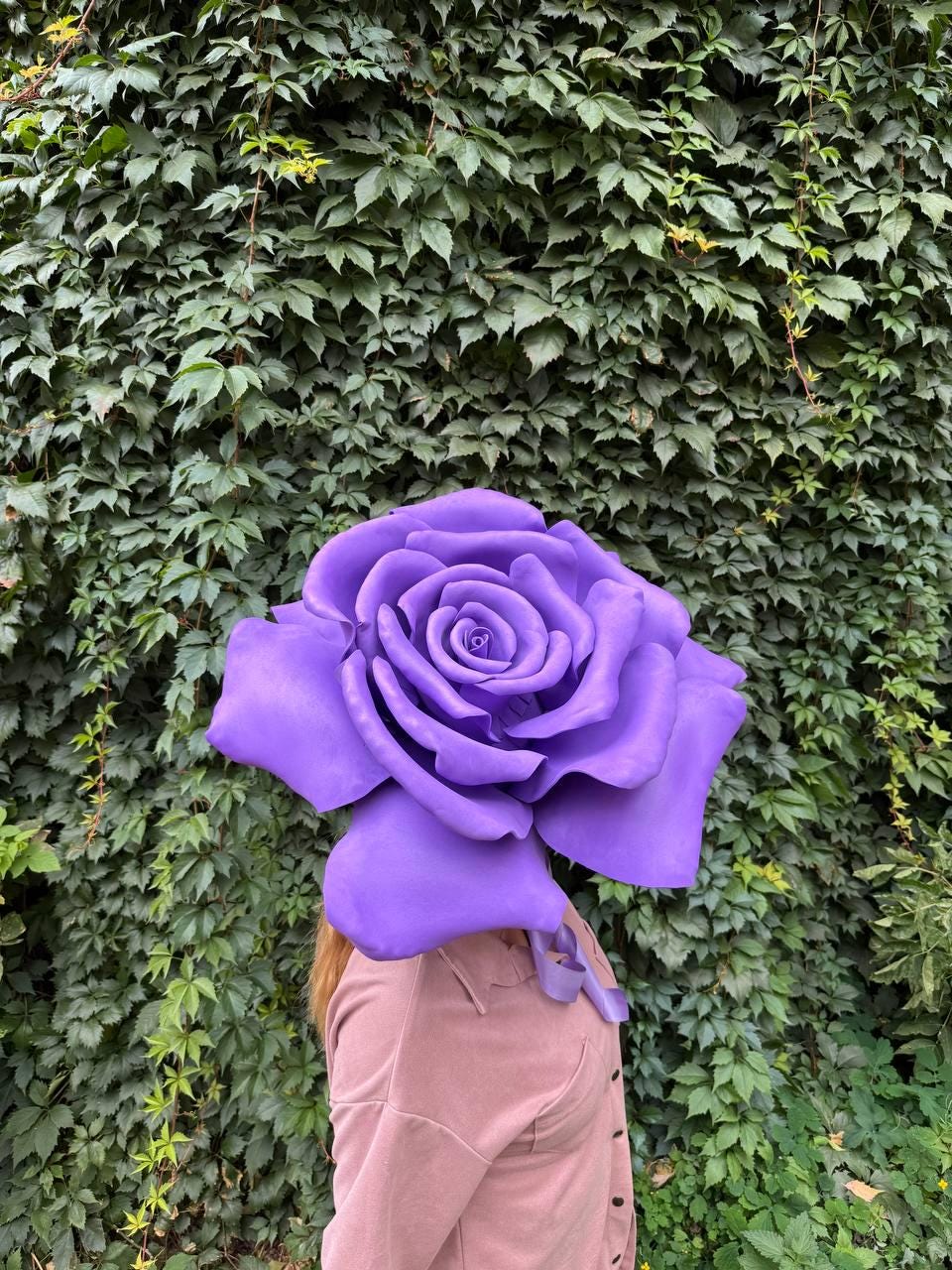 May include: A large, vibrant purple rose headpiece. The oversized flower is the focal point, with detailed petals and a deep purple hue. The background is a lush green wall of foliage. The headpiece is worn by a person wearing a pink shirt.