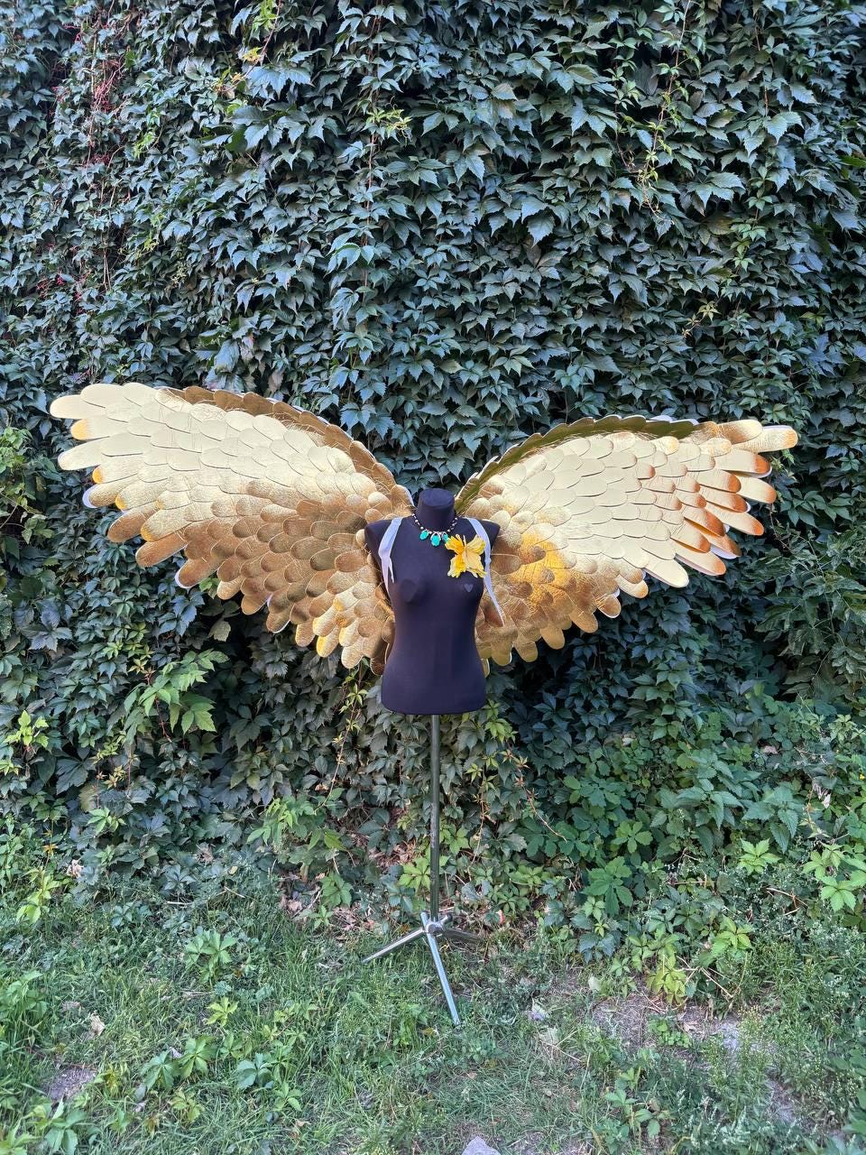 May include: Gold-colored costume wings with layered feather design, displayed on a black mannequin. The wings are detailed with shades of gold and brown, and the mannequin is adorned with a necklace and a yellow flower. The background features lush green foliage.