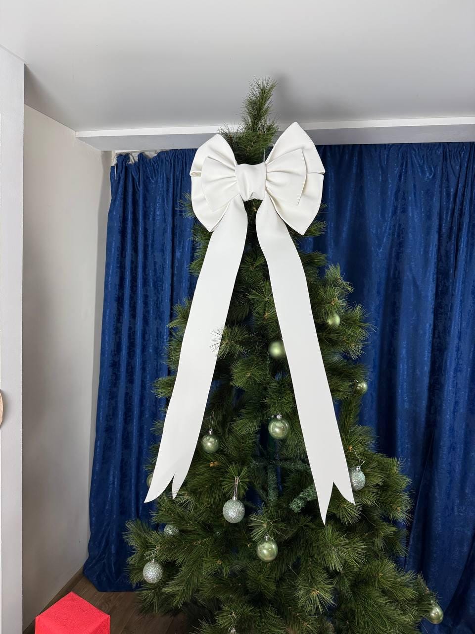 May include: A white bow Christmas tree topper with long, flowing ribbons. The bow is attached to a green Christmas tree decorated with silver and green ornaments. The background features a blue curtain.