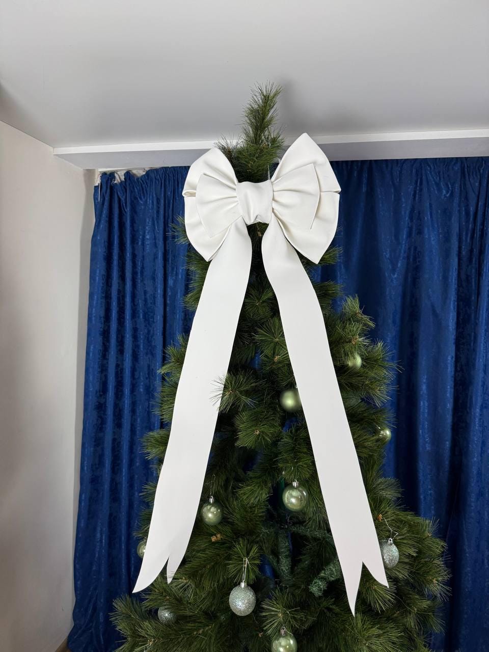 May include: A large, white bow tree topper with long, flowing ribbons adorns a green Christmas tree. The bow is centered at the top of the tree, with the ribbons cascading down. The tree is decorated with green and silver ornaments.