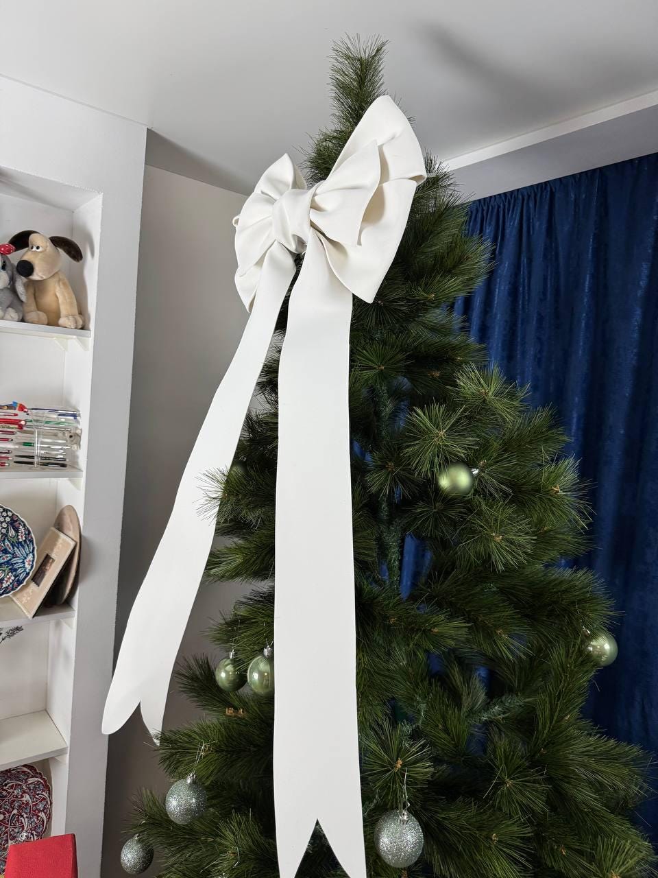 May include: A large, off-white bow adorns the top of a green Christmas tree. The bow has long, flowing tails. The tree is decorated with silver and green ornaments. The background includes a white wall, a shelf with toys, and blue curtains.