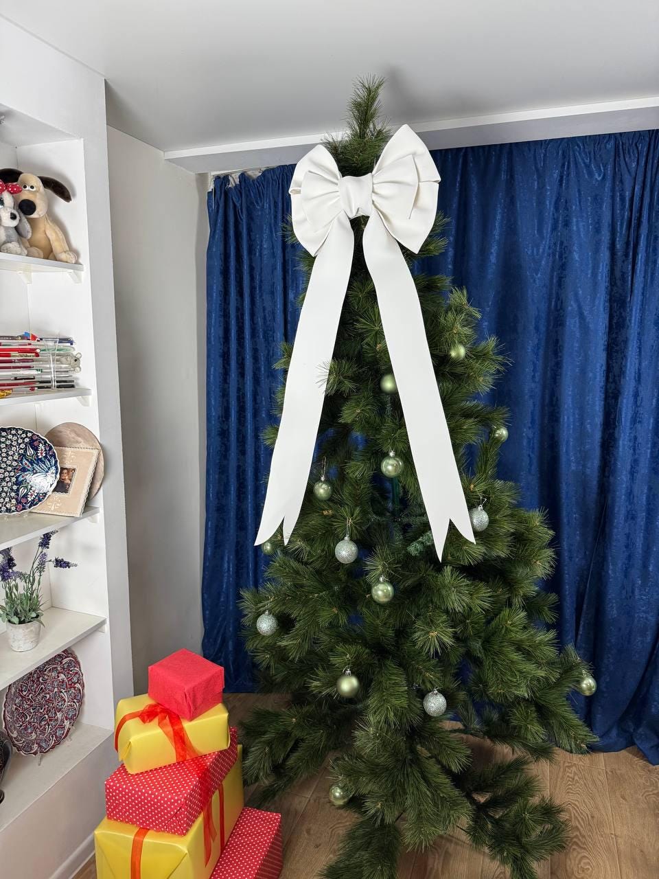 May include: A decorated Christmas tree with a large white bow at the top and silver ornaments. Several wrapped gifts in red and yellow are stacked at the base of the tree. The background features blue curtains.