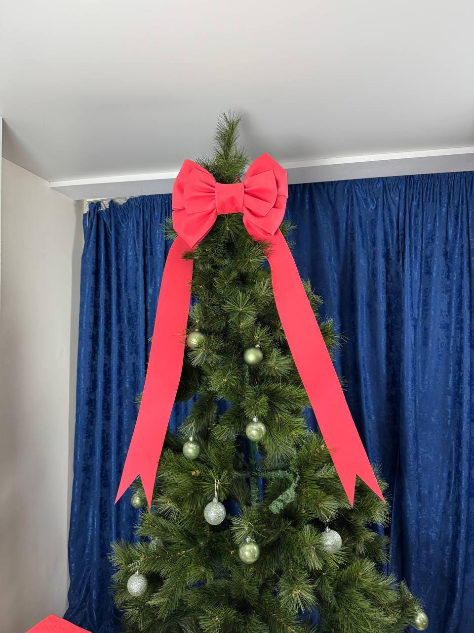 May include: A decorated Christmas tree with a large red bow as a tree topper. The tree is adorned with green and silver ornaments. The bow's long ribbons cascade down the tree, creating a festive holiday display.