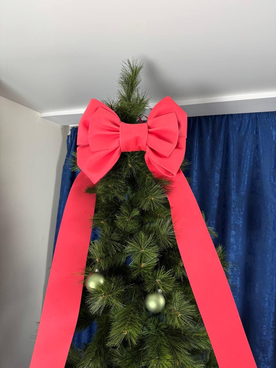 May include: A Christmas tree topped with a large, bright red bow. The bow is made of fabric and has long, flowing tails. Two green ornaments hang from the tree's branches.