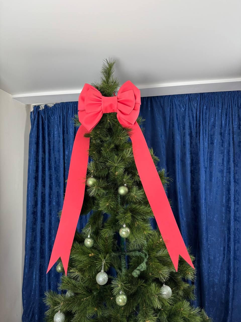May include: A decorated Christmas tree with a large red bow at the top and long red ribbons cascading down. The tree is adorned with silver and green ornaments, set against a blue curtain backdrop.