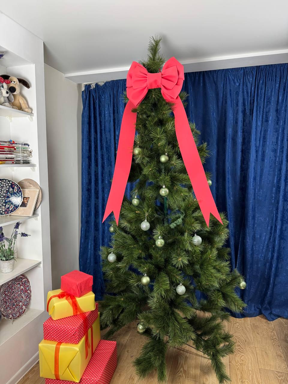 May include: A decorated Christmas tree with a large red bow at the top and ornaments. Several wrapped gifts in red and yellow are stacked at the base of the tree. The tree is set against a blue curtain backdrop.