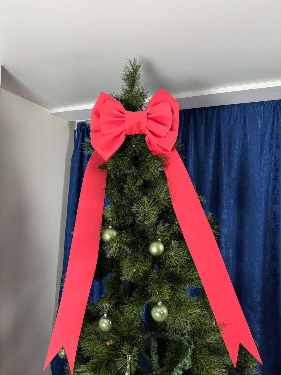 May include: A decorated Christmas tree with a large red bow at the top and long, flowing ribbons. The tree is adorned with green ornaments. The background includes a blue curtain and a white ceiling.