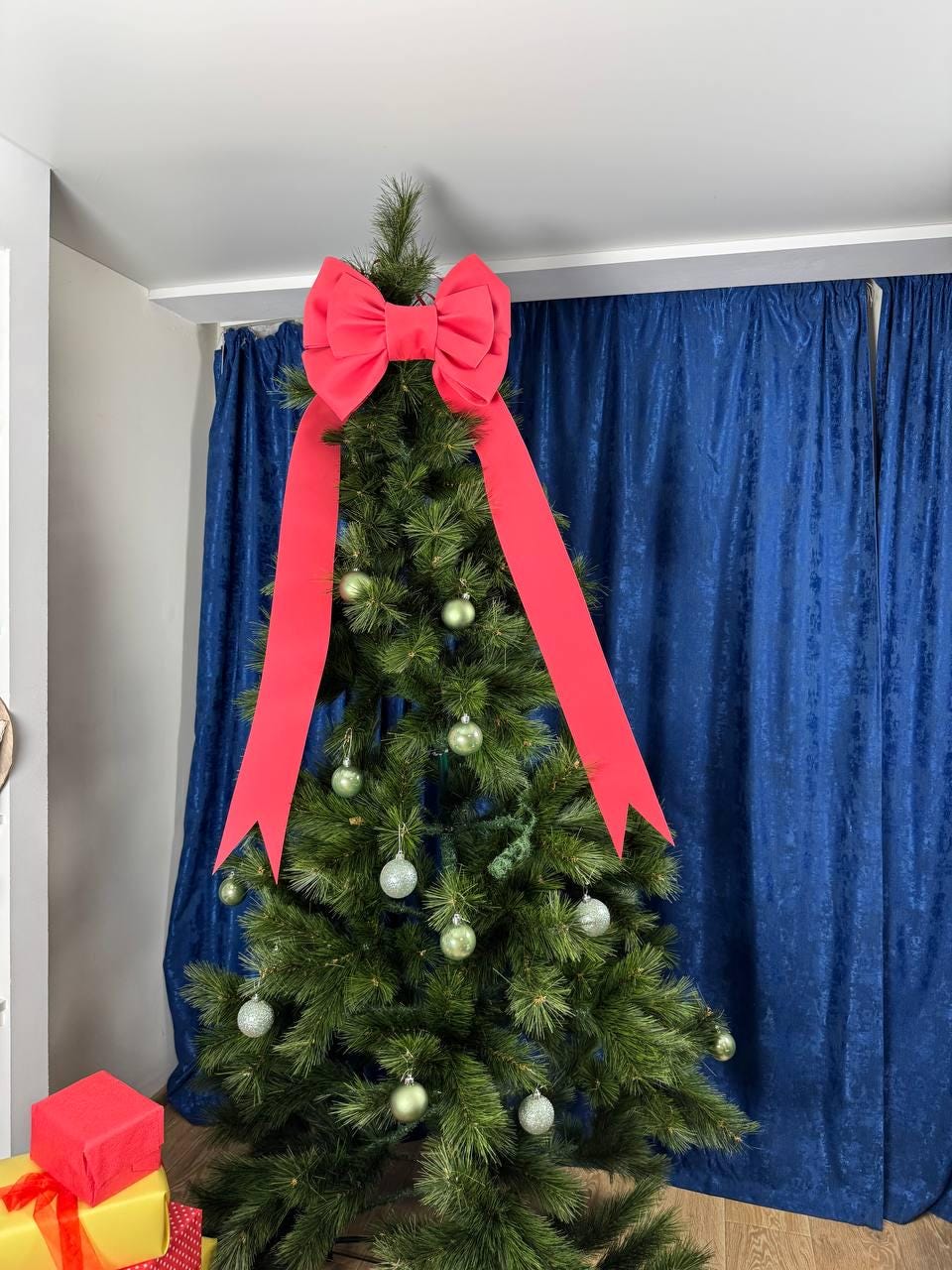 May include: A decorated Christmas tree with a large red bow at the top and long flowing ribbons. The tree is adorned with silver and green ornaments. A red gift box is visible in the lower left corner, suggesting a festive holiday theme.