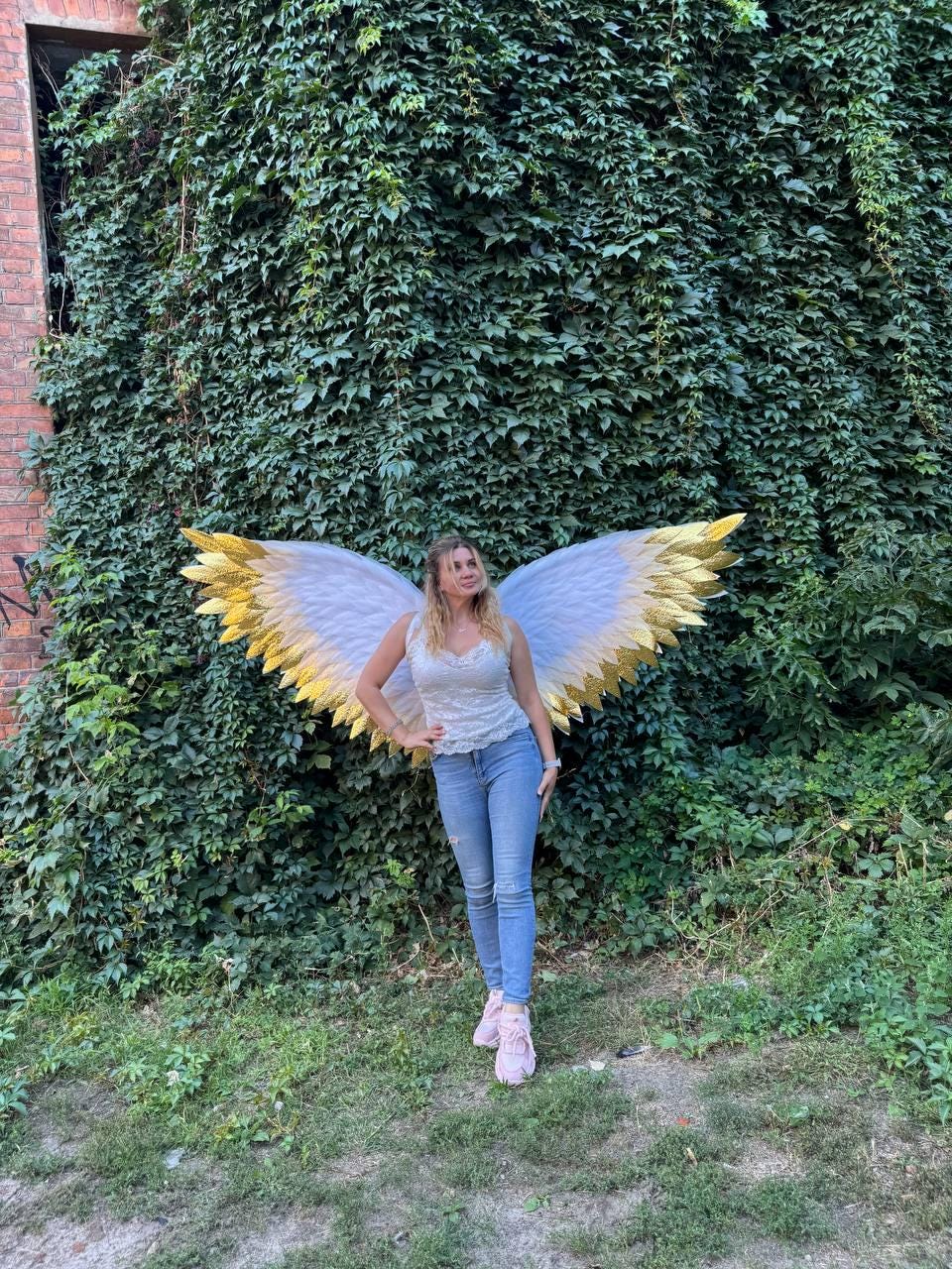 May include: A person wearing white and gold angel wings, standing in front of a wall covered in green vines. The wings have white feathers with gold tips. The person is wearing a white top, blue jeans, and pink sneakers.