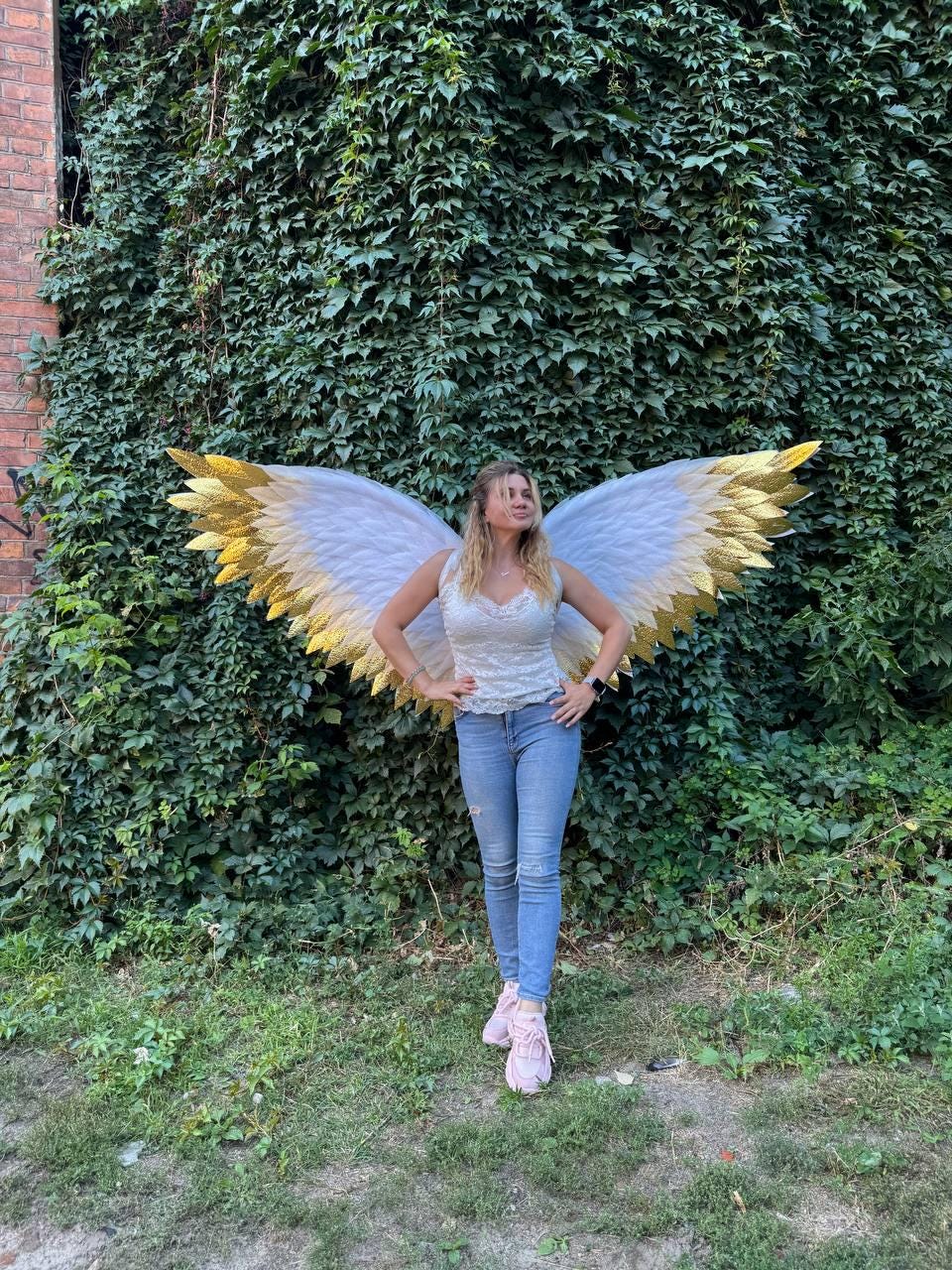 May include: White and gold angel wings with a woman wearing a white top and blue jeans. The wings have white feathers with gold tips. The woman is standing in front of a wall of green foliage.