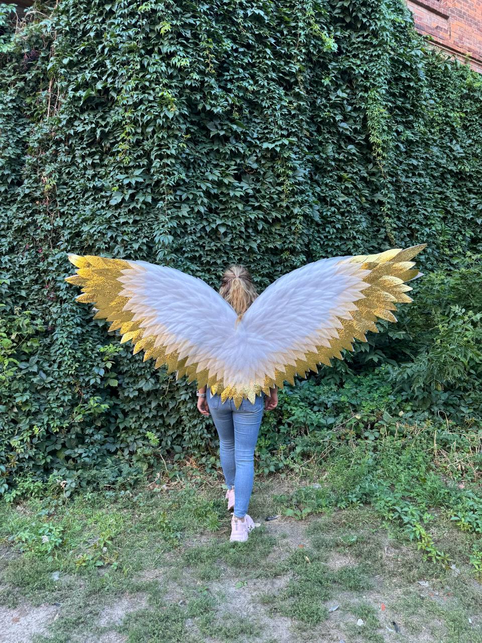 May include: White and gold glitter angel wings with a feathered design. The wings are spread wide, showcasing the intricate details and textures. The wings are worn by a person in blue jeans and pink sneakers, standing in front of a wall covered in green foliage.