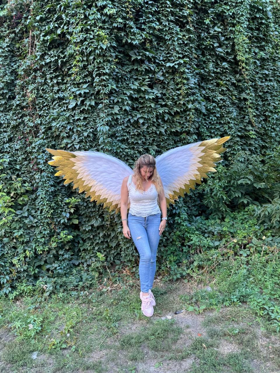 May include: A person wearing white and gold angel wings stands in front of a wall of green foliage. The wings have white feathers with gold tips. The person is wearing a white top, blue jeans, and pink sneakers.