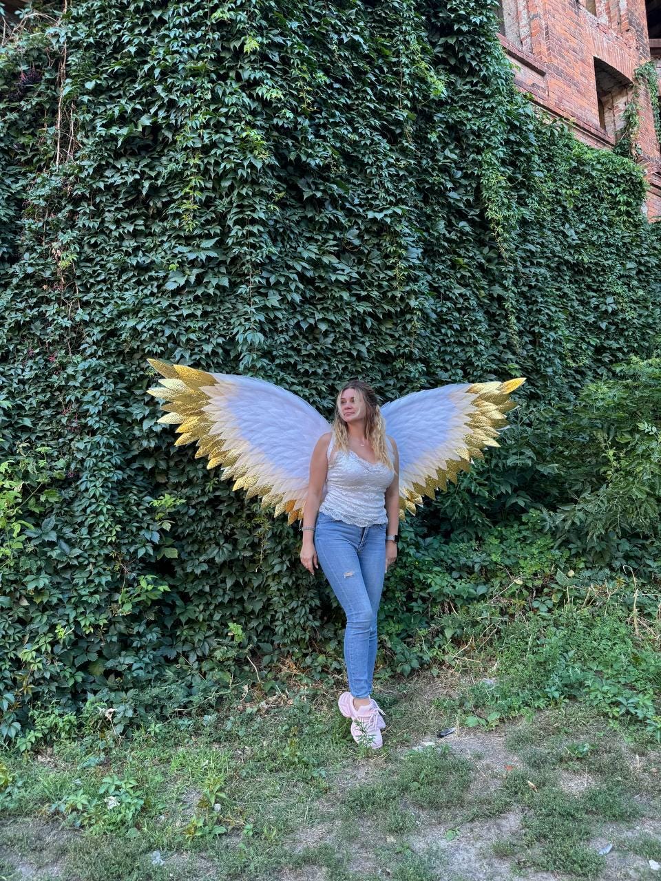 May include: A person wearing white and gold angel wings stands in front of a wall of green ivy. The wings have white feathers with gold tips. The person is wearing a white top, blue jeans, and pink shoes.