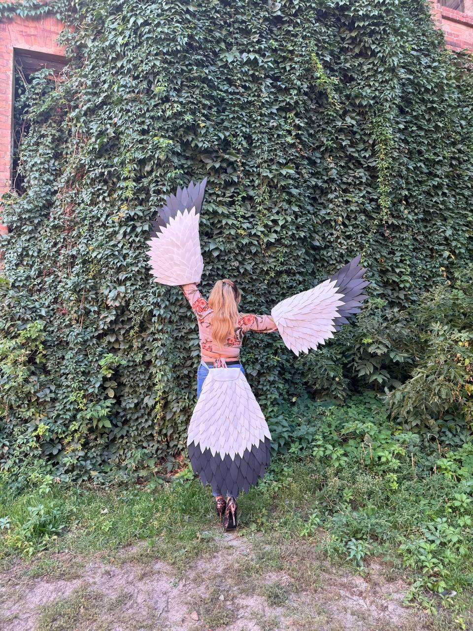 May include: A person wearing a costume with large, white, gray, and black feathered wings and a matching tail. The costume is set against a backdrop of lush green foliage and a brick building.