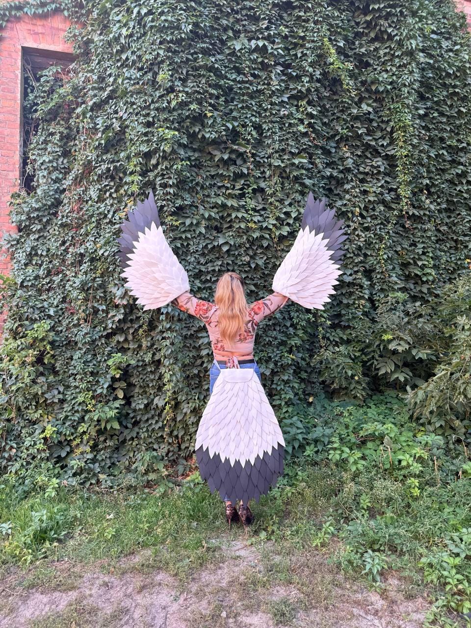 May include: A person wearing a costume with large, white, gray, and black feathered wings and tail. The costume is made of layered, geometric shapes. The person is standing in front of a wall of green foliage.