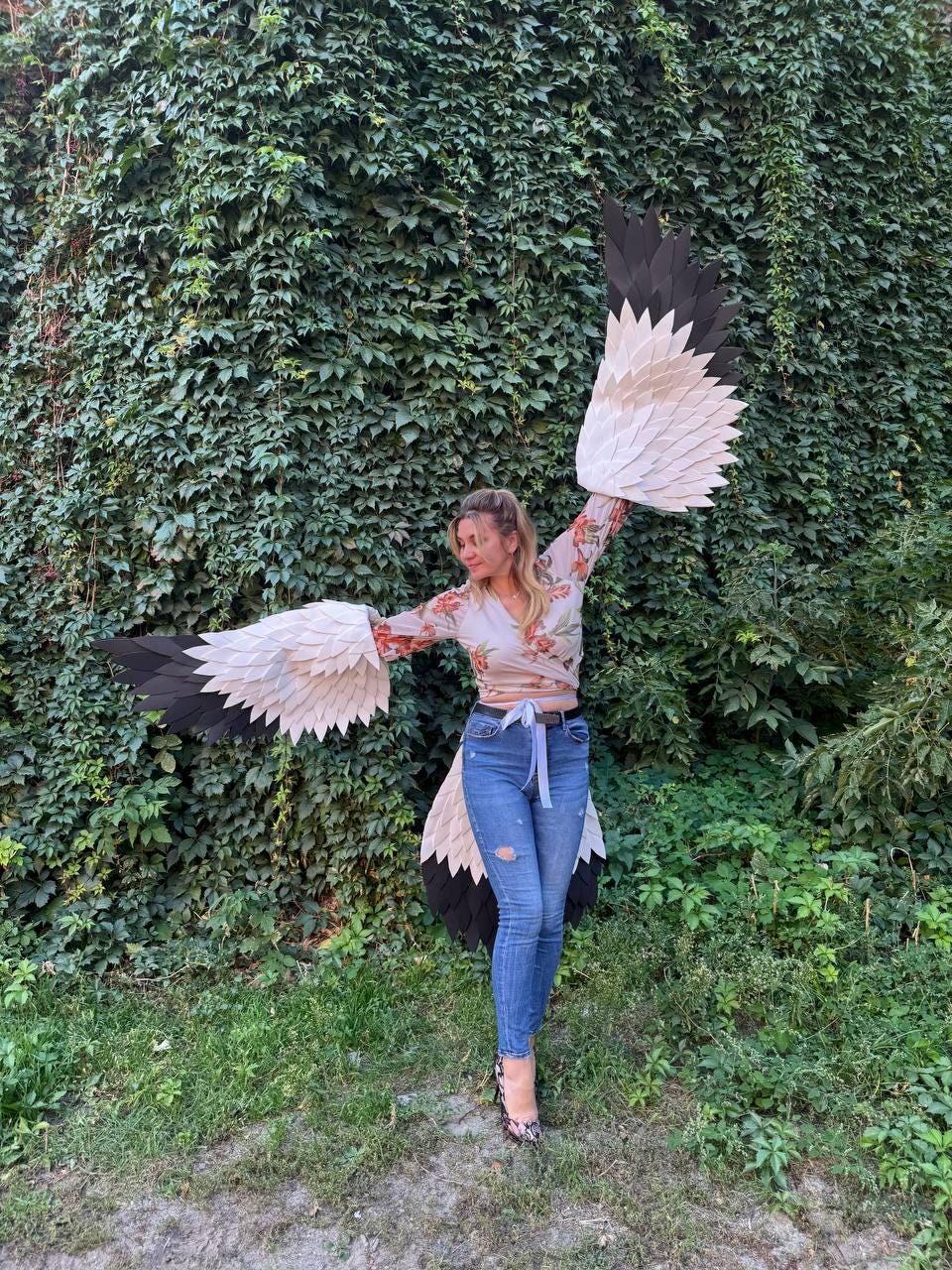 May include: A person wearing large, handmade bird wings with black, white, and tan feathers. The wings are spread wide against a backdrop of green foliage. The person is wearing a floral top and blue jeans.