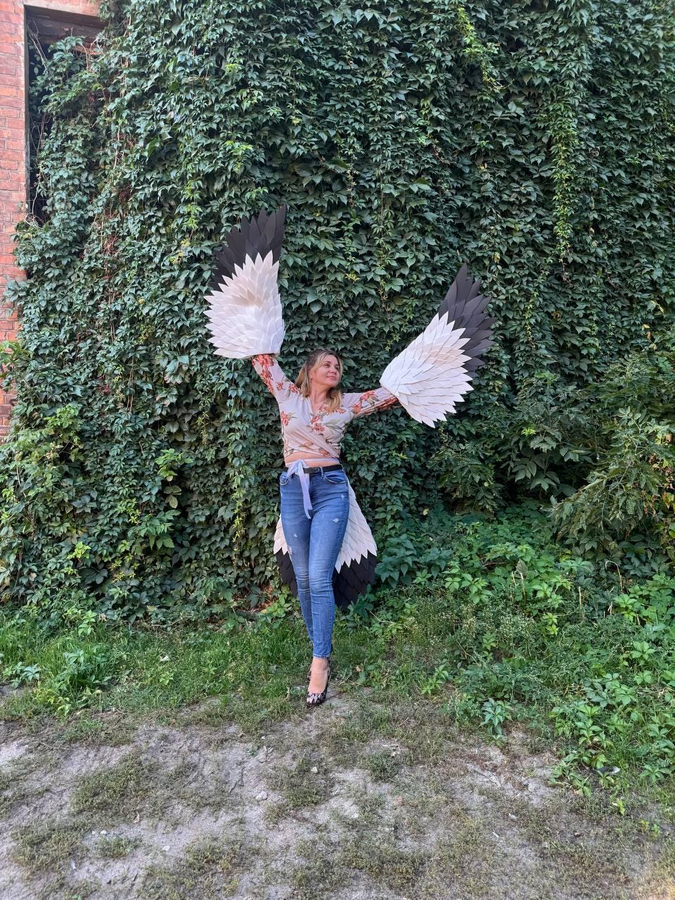 May include: A person wearing a floral top and blue jeans, posing with large, handmade bird wings. The wings feature white, black, and beige feathers, creating a striking visual contrast against the green foliage background.