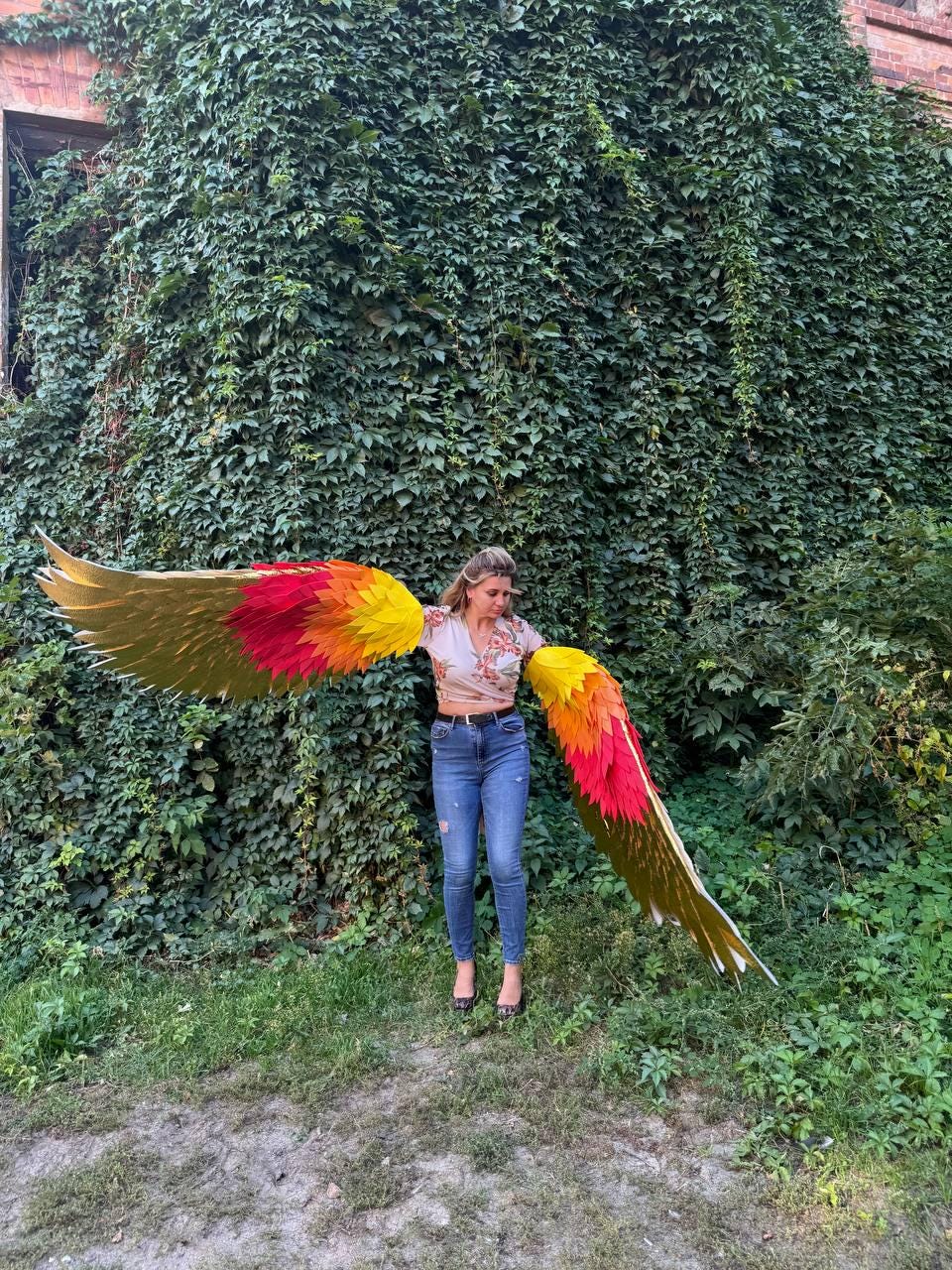 May include: Large, colorful wings with gold, red, orange, and yellow feathers. The wings are spread wide, creating a striking visual against a backdrop of lush green foliage. The wings appear to be costume or decorative items, possibly for cosplay or theatrical use.