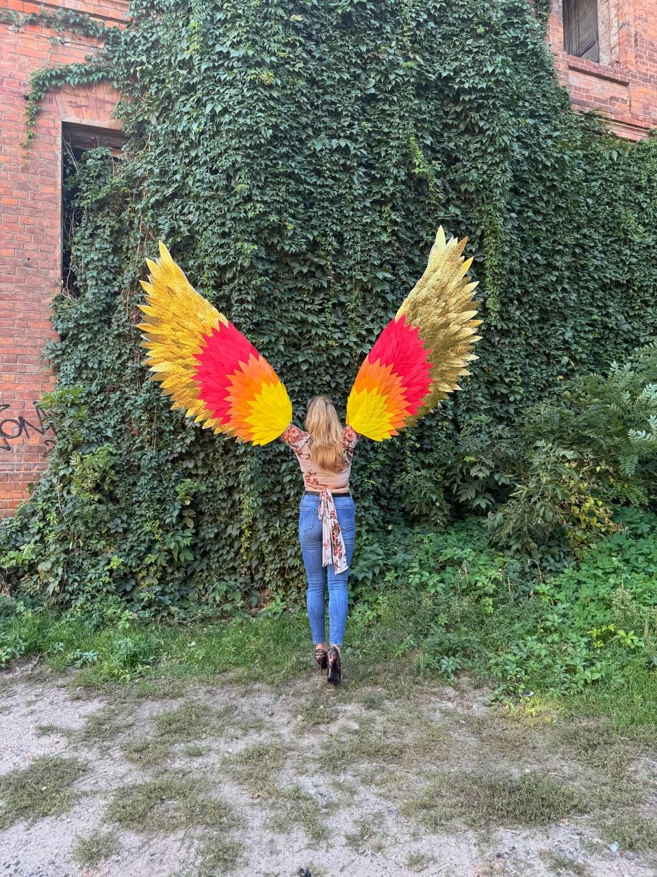 May include: A person wearing colorful, large, decorative wings with gold, yellow, orange, and red hues, standing in front of a wall covered in green foliage. The wings are spread wide, creating a striking visual effect.