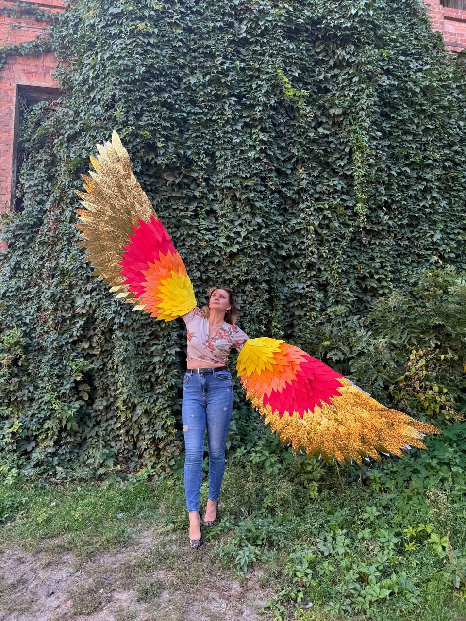 May include: A person wearing large, colorful, feathered wings. The wings feature gold, red, orange, and yellow hues. The person is standing in front of a wall covered in green foliage.