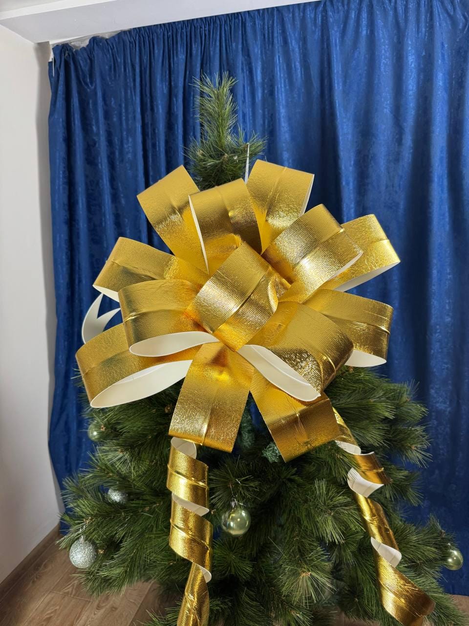 May include: A gold metallic bow sits atop a small Christmas tree. The bow is made of shiny, textured material and has a large, multi-layered design. Green ornaments and curly ribbons add to the festive look. The tree is set against a blue backdrop.