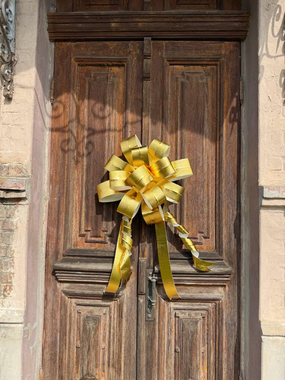 May include: A large, golden-yellow bow adorns a weathered, dark brown wooden door. The bow is made of shiny, metallic ribbon and is tied in a decorative fashion. The door has a classic design with panels and a metal handle.