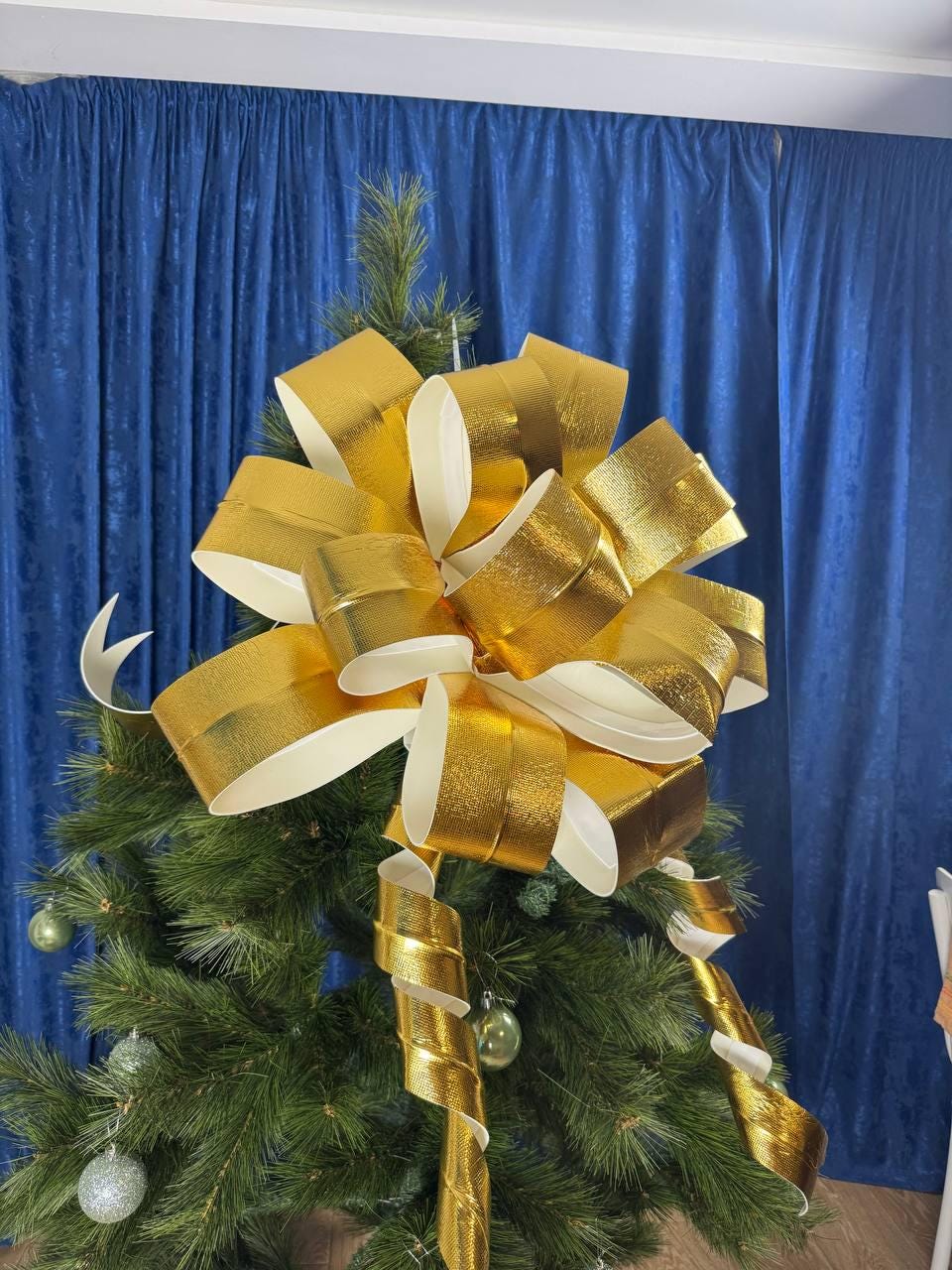 May include: A gold and white Christmas tree bow adorns a green artificial tree. The bow is made of shiny, metallic gold ribbon with white undersides. Gold ribbon spirals cascade down the tree, and silver and green ornaments are visible.