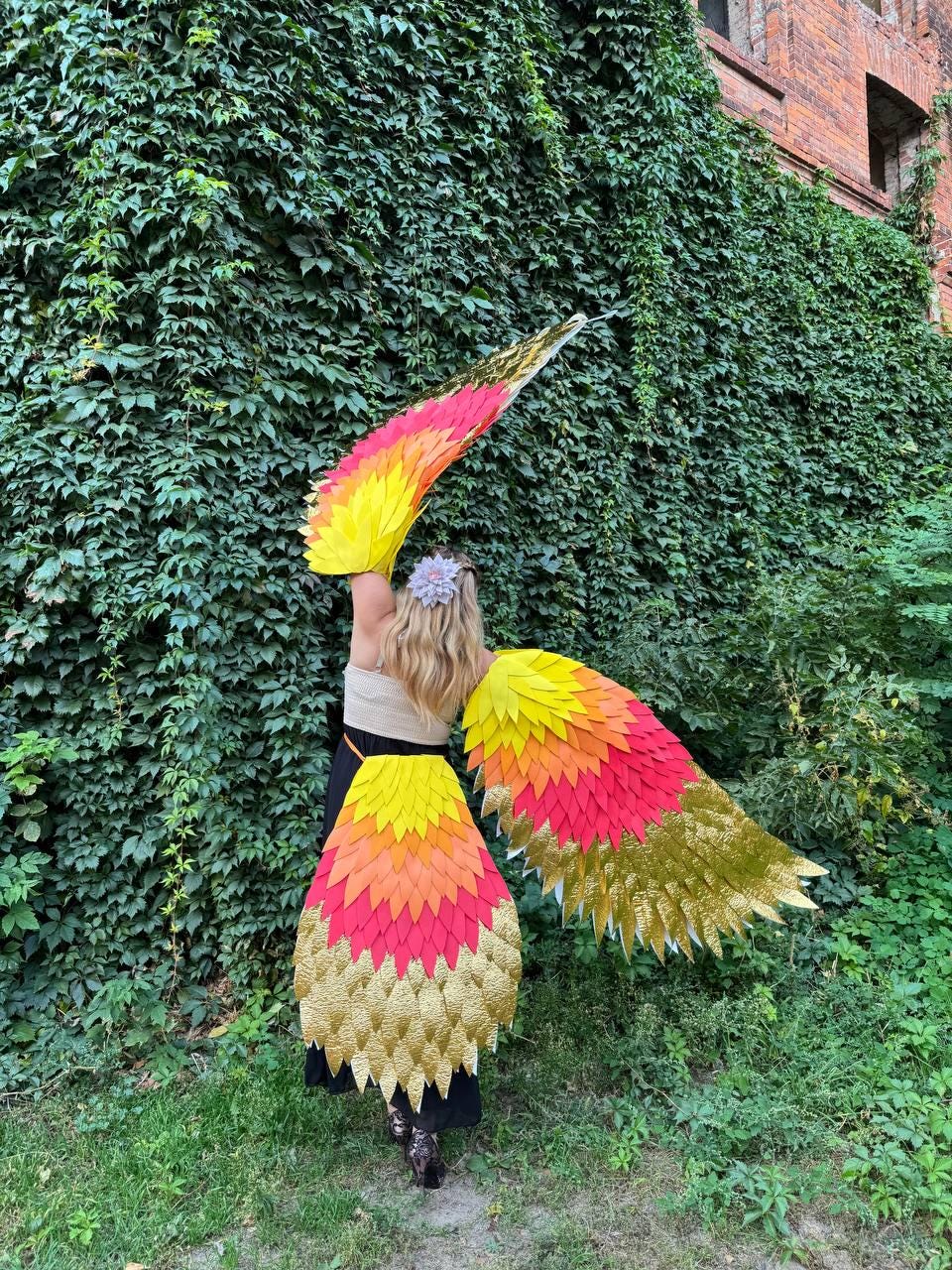 May include: A person wearing colorful, layered costume wings. The wings feature a gradient of yellow, orange, and red, transitioning to gold at the tips. The costume is set against a backdrop of green foliage and a brick building.