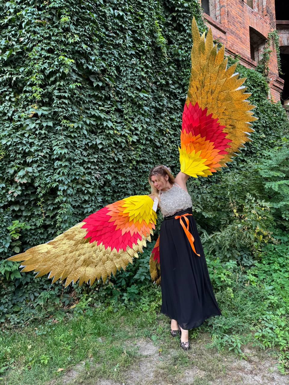 May include: Large, colorful costume wings with gold, red, orange, and yellow hues. The wings are spread wide, showcasing the vibrant gradient of colors. The wings are the focal point, with a person wearing them, dressed in a black skirt and silver top.