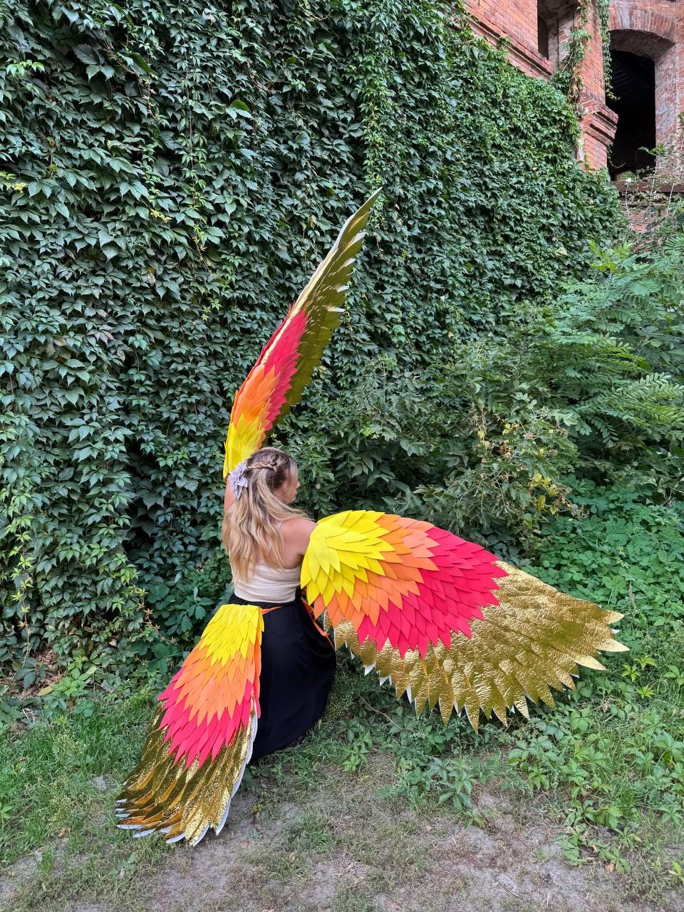 May include: A person wearing large, colorful costume wings. The wings feature layers of yellow, orange, and red, with gold accents. The person is wearing a cream-colored top and black skirt, posing outdoors.