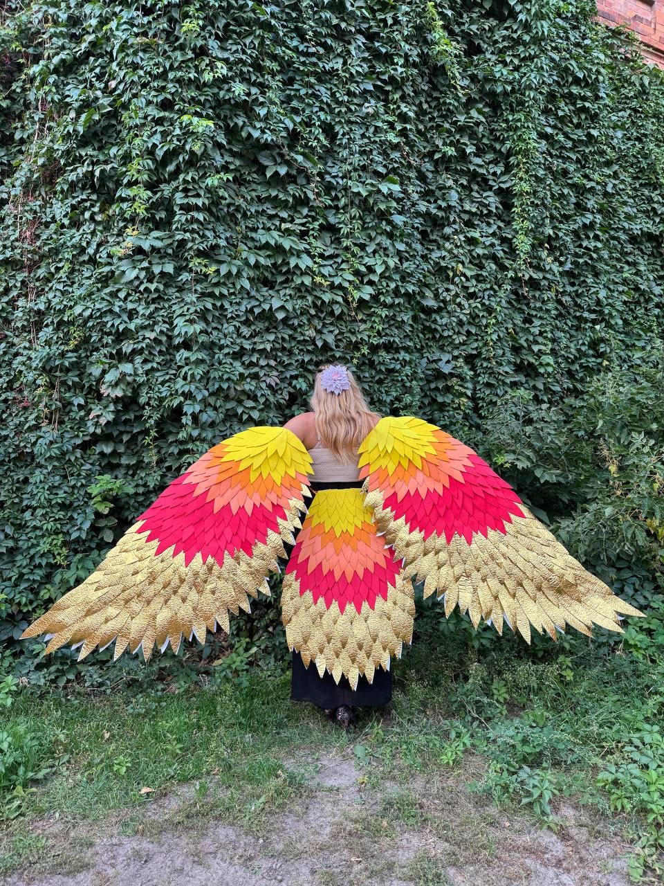 May include: A person wearing large, colorful, feathered wings. The wings transition from gold to red, orange, and yellow. The person is standing in front of a wall of green foliage.