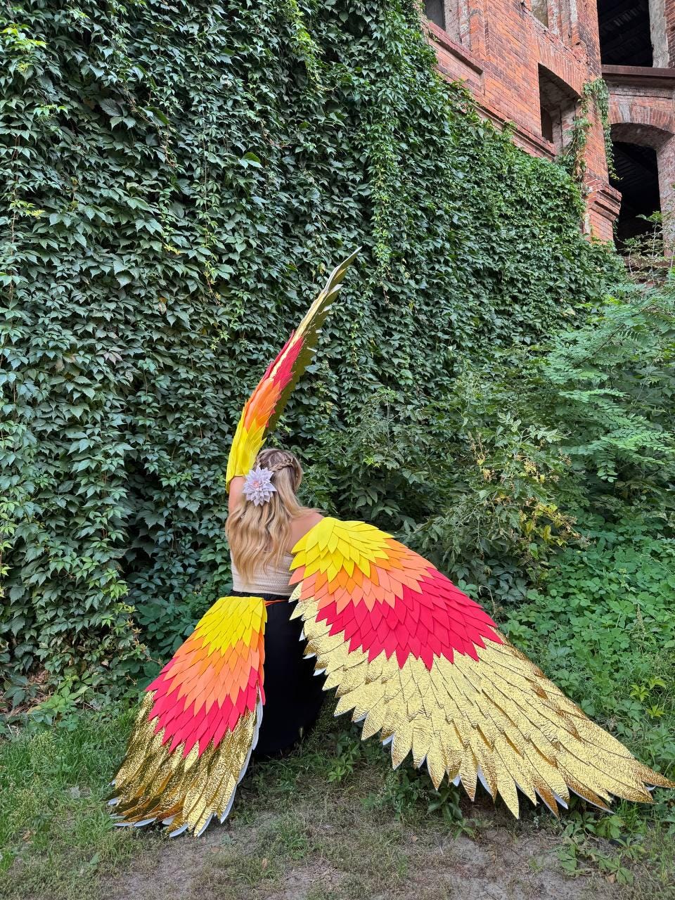 May include: A pair of large, colorful wings with a gradient of red, orange, and yellow feathers, edged with gold. The wings are spread against a backdrop of lush green foliage and a brick building.