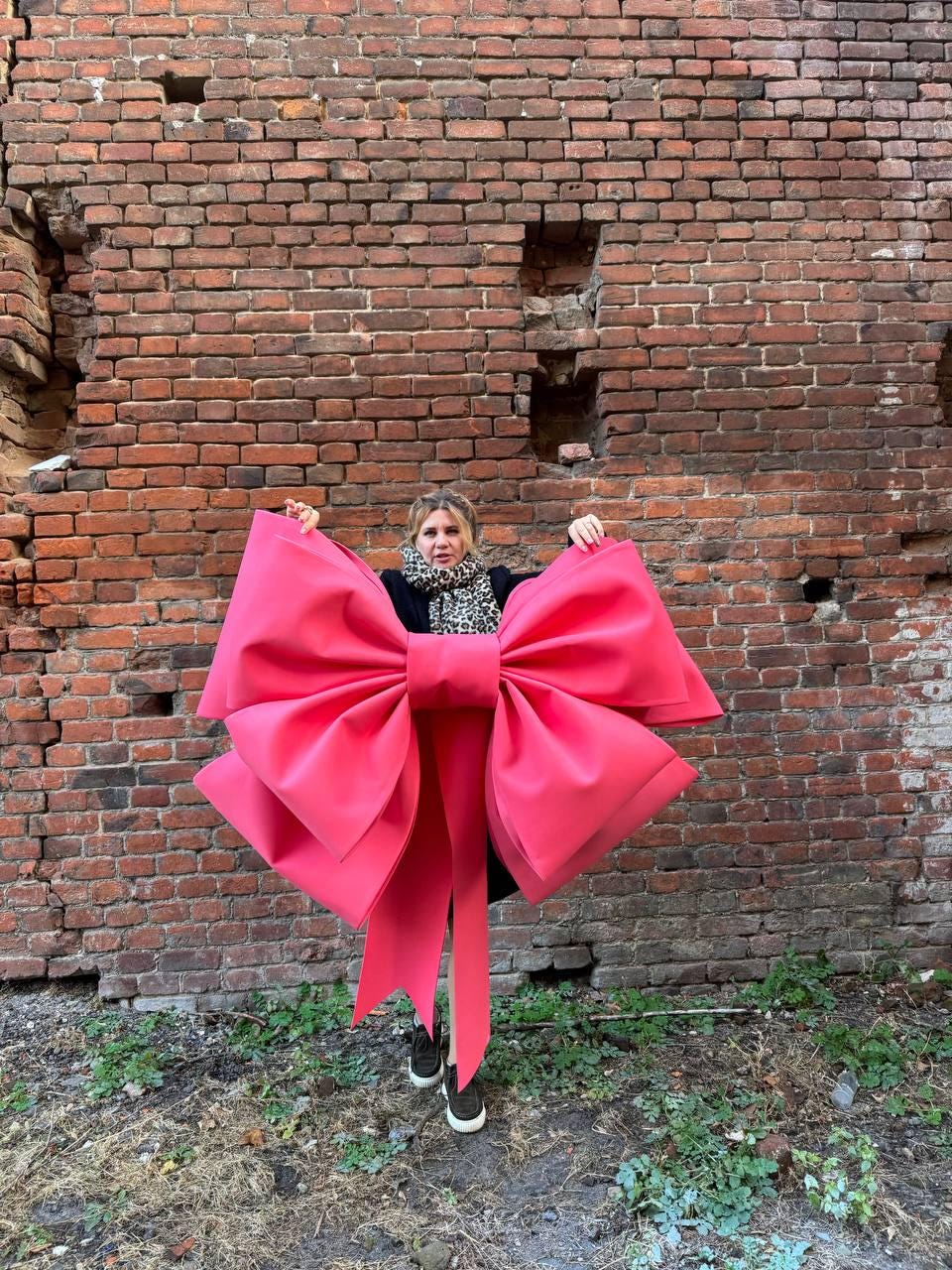 May include: A large, vibrant pink bow with long, flowing tails. The bow is held up against a weathered brick wall, creating a striking contrast. The bow's size suggests it could be used for decoration or as a prop.