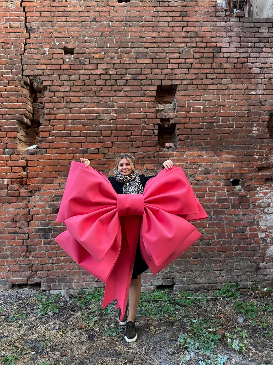 May include: A large, vibrant pink bow with long streamers. The bow is held up against a weathered brick wall. The bow's size and color make it a striking decorative element, suitable for events or displays.