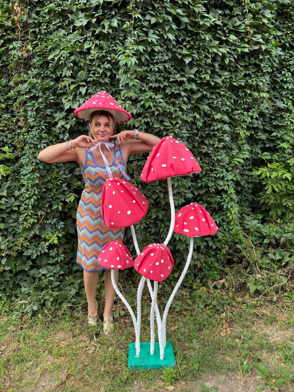 May include: A whimsical display featuring a cluster of large, red mushroom decorations with white polka dots. The mushrooms are supported by white stems and a green base. A woman is wearing a matching mushroom hat and a colorful dress.