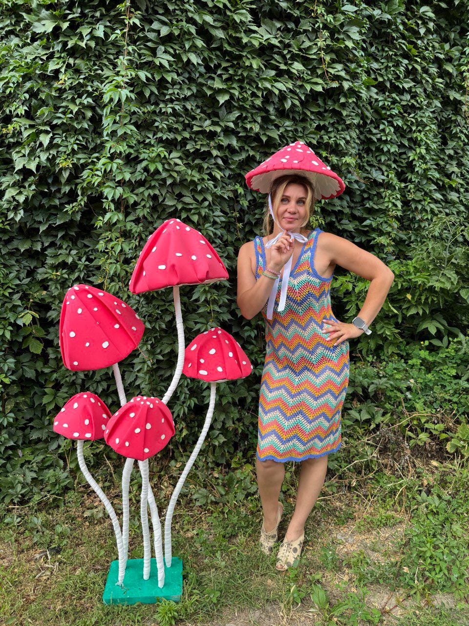 May include: A whimsical display featuring a cluster of red mushroom sculptures with white polka dots, set against a backdrop of lush greenery. The mushrooms have white stems and are mounted on a green base. A woman is wearing a matching mushroom hat and a colorful crochet dress.