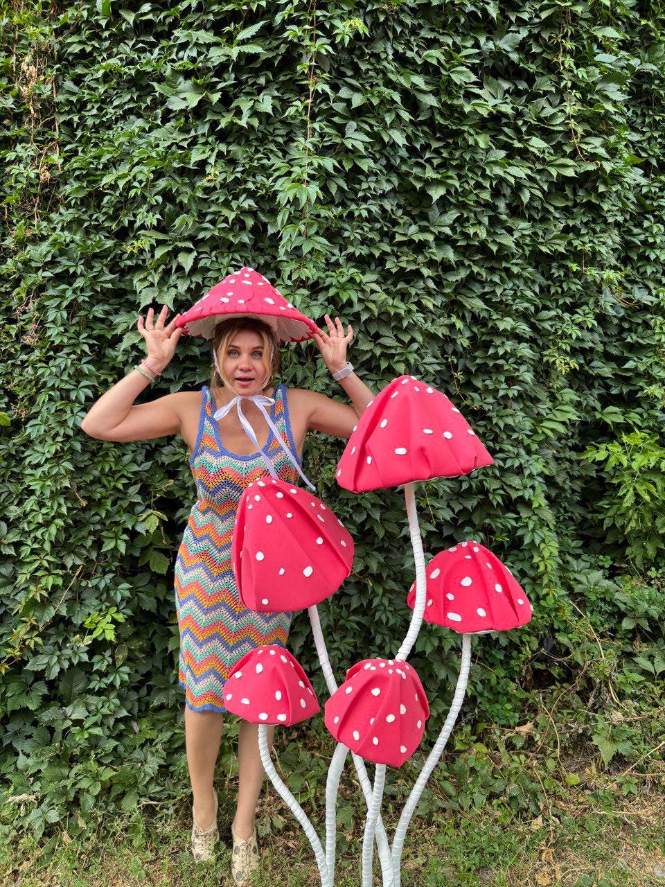 May include: A woman wearing a red mushroom hat and a colorful crochet dress stands in front of a green leafy background. Several large, red mushroom decorations with white polka dots are positioned nearby, creating a whimsical scene.