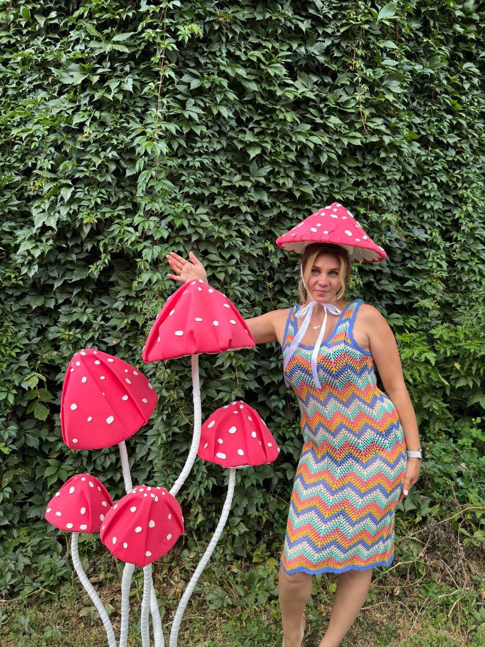 May include: A woman wearing a colorful crochet dress and a mushroom-shaped hat poses next to a cluster of large, red mushroom decorations with white polka dots. The mushrooms have white stems and are set against a backdrop of green foliage.