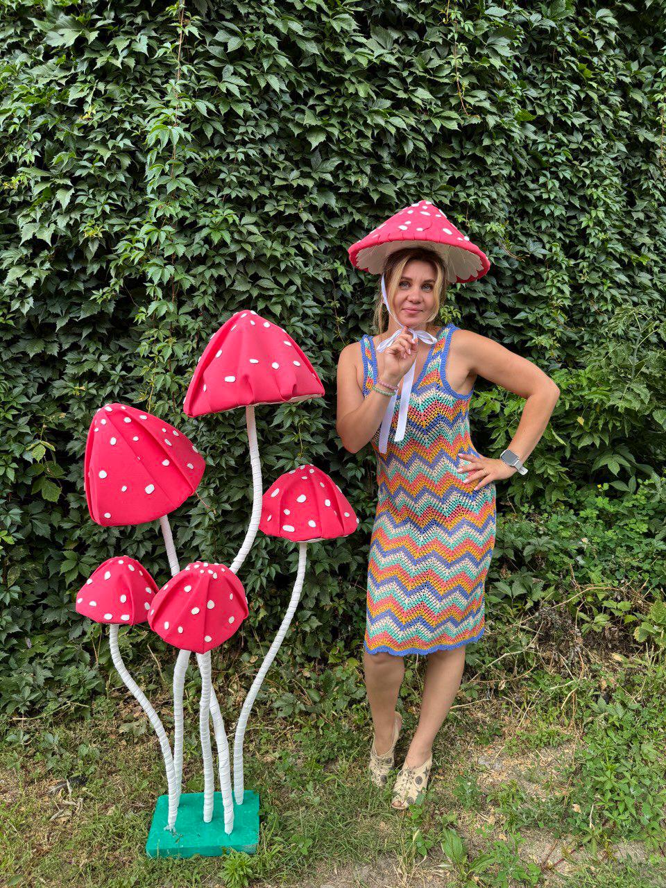 May include: A whimsical display featuring a cluster of red mushroom sculptures with white polka dots and a matching mushroom hat. The mushrooms have white stems and are set on a green base. A colorful crochet dress completes the playful theme.