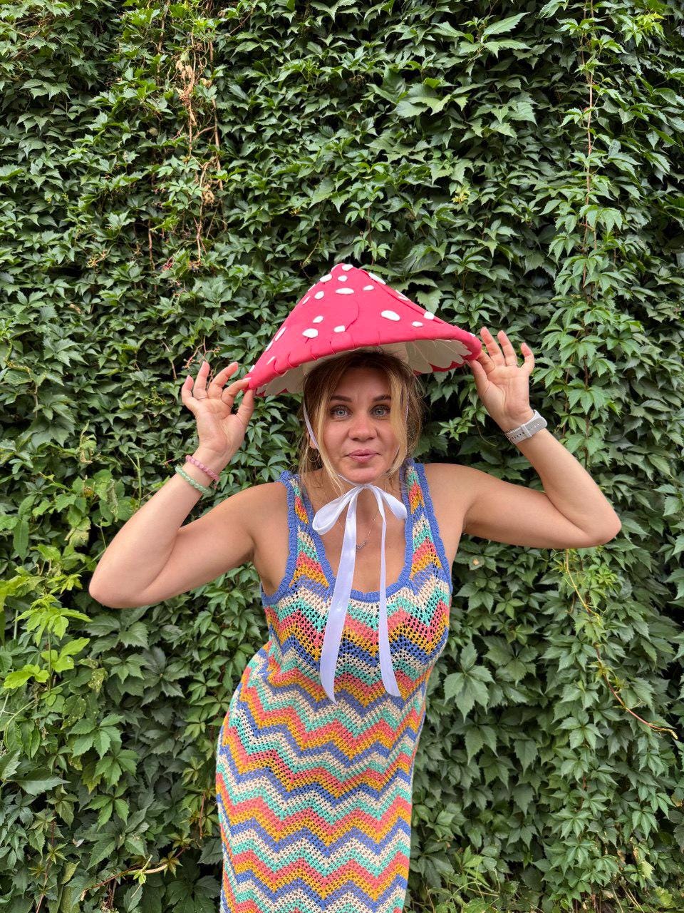 May include: A red mushroom-shaped hat with white polka dots is held up by a person. The person is wearing a colorful, sleeveless, chevron-patterned dress with a white ribbon. The background is a wall of green leaves.