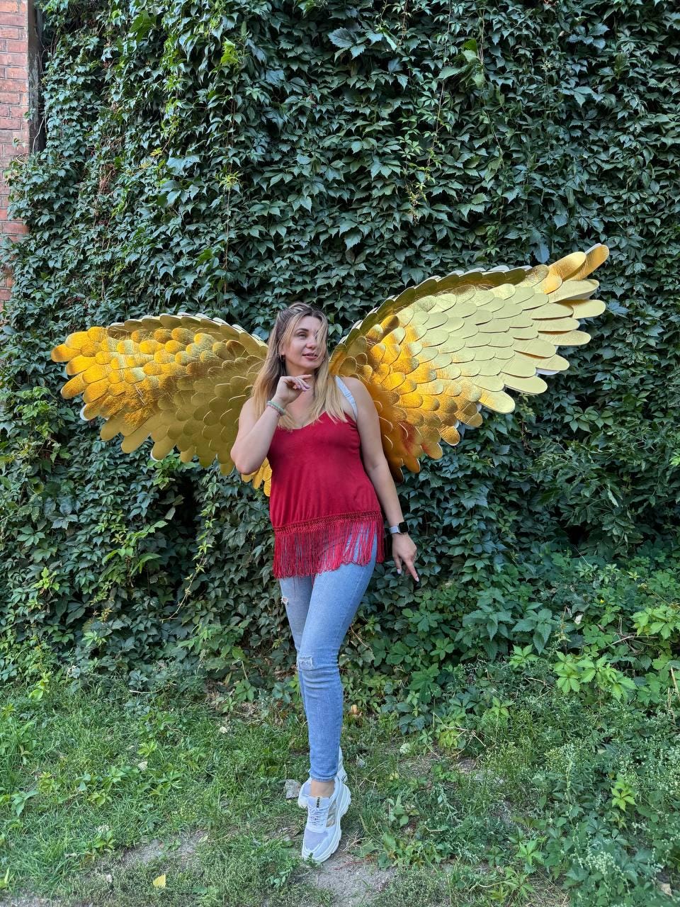 May include: A person wearing large, golden angel wings with a layered feather design. The wings are the focal point, set against a backdrop of lush green foliage. The person is wearing a red top and blue jeans.